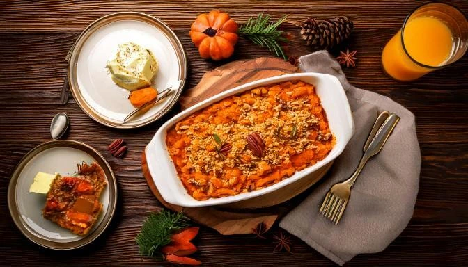 A baked sweet potato casserole with a pecan streusel topping, served in a white dish alongside plated portions, butter, and festive fall decorations.